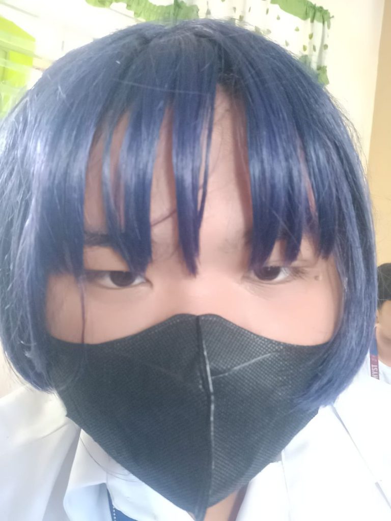 A person with short, dark blue hair is wearing a black face mask. The background appears to be indoors with a green and white curtain partially visible. The person is looking directly at the camera with a neutral expression.