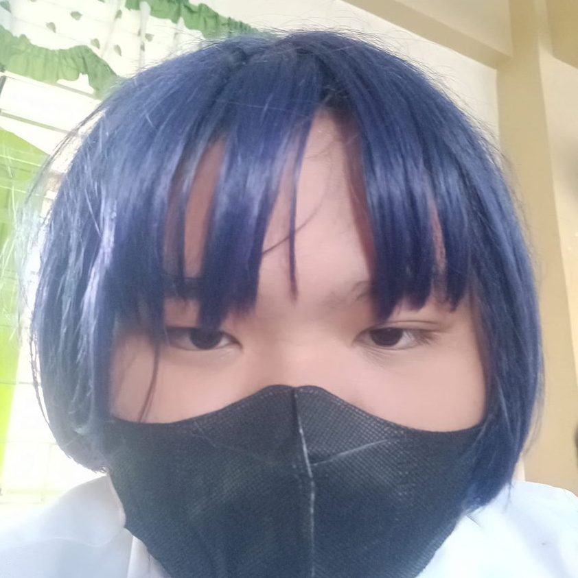 A person with blue hair and bangs is wearing a black face mask. They are indoors, with green and white curtains visible in the background.